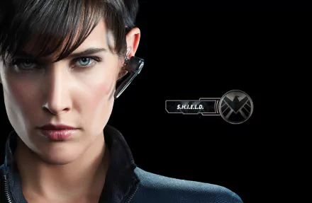 A close-up of a confident character from Marvel's Agents of S.H.I.E.L.D., featuring a headset and S.H.I.E.L.D. emblem against a sleek black background.
