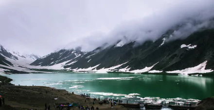 Snow-covered mountain landscape in Pakistan with a partially frozen lake, scattered ice, clouds hovering above, and people gathered along the shore in this HD nature photography.