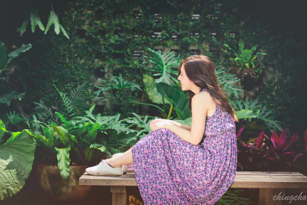 A Taiwanese model sits gracefully on a bench in a lush garden, wearing a floral dress. The serene backdrop enhances the beauty of the scene. 4K Ultra HD wallpaper.
