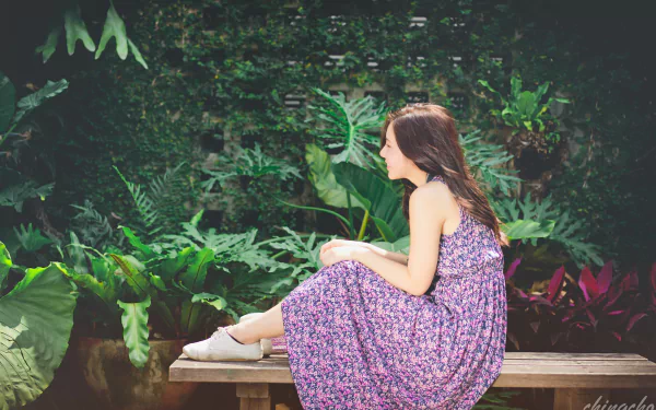 A Taiwanese model sits gracefully on a bench in a lush garden, wearing a floral dress. The serene backdrop enhances the beauty of the scene. 4K Ultra HD wallpaper.