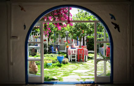 A vibrant garden view seen through an arched doorway, featuring colorful flowers, lush greenery, and a welcoming dining area, captured in a stunning HD wallpaper.