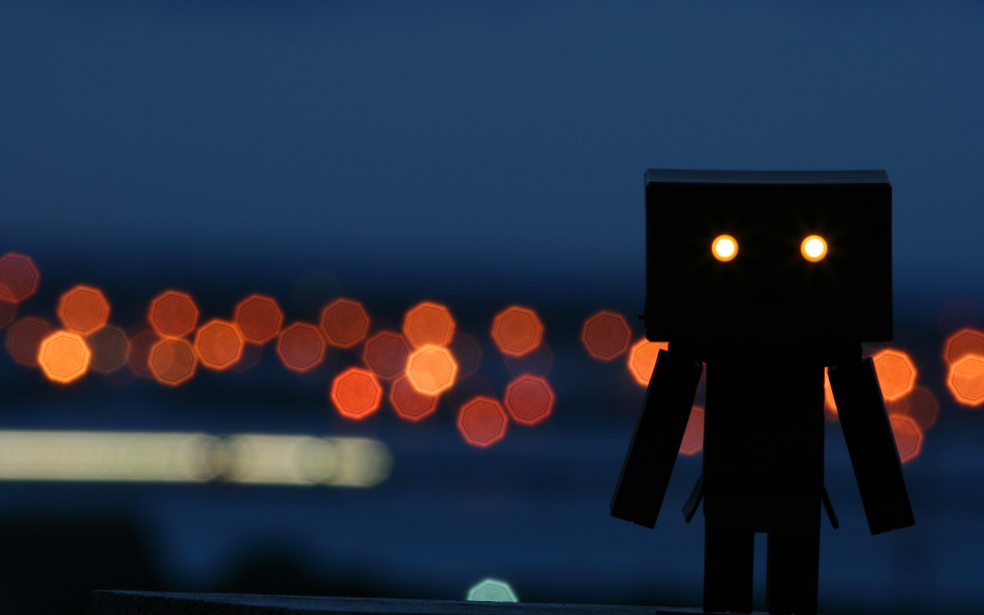 HD PC desktop wallpaper featuring a Danbo figure with glowing eyes silhouetted against a blurred cityscape of warm bokeh lights at dusk.