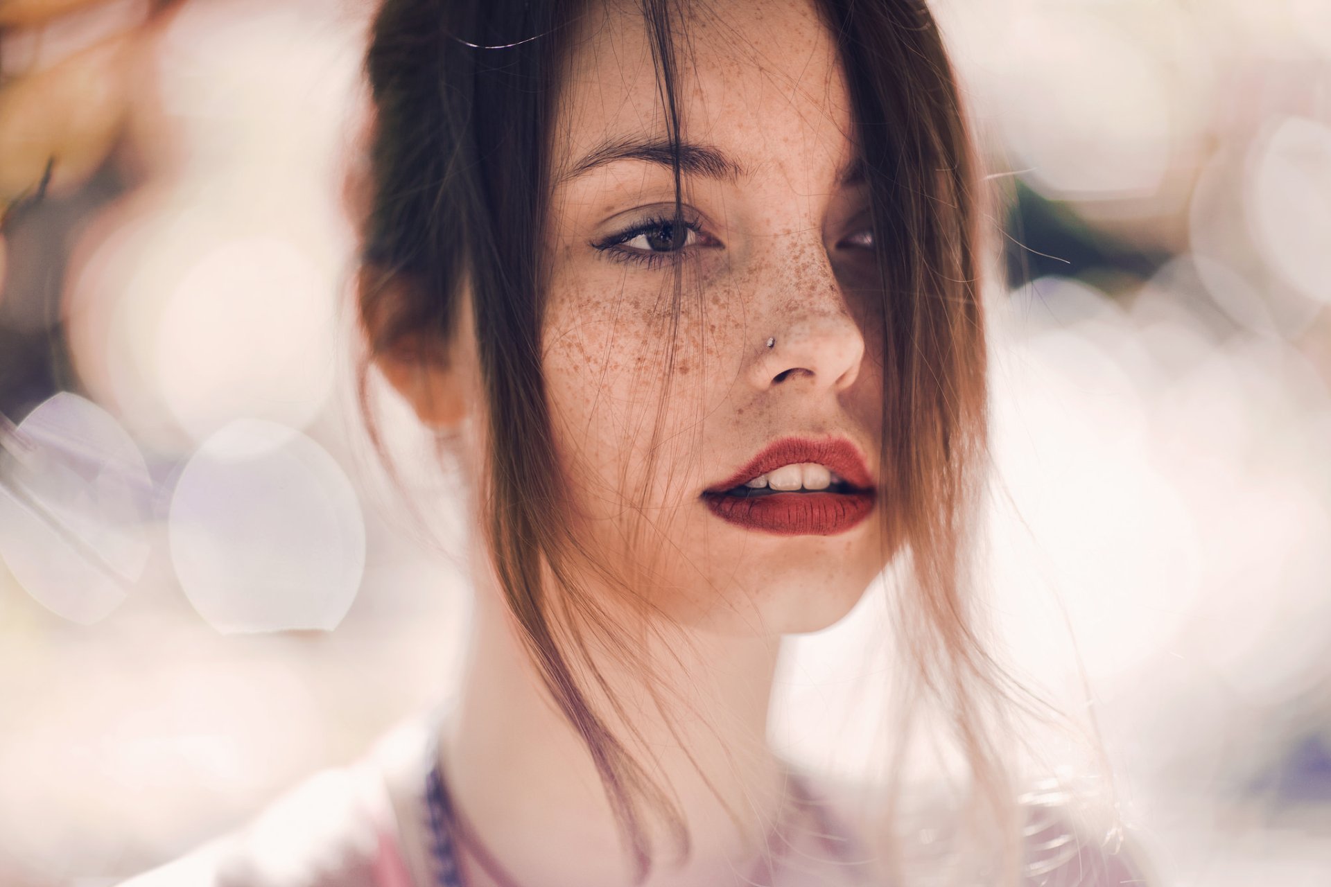 A brunette woman with freckled skin and bold red lipstick gazes confidently, set against a soft bokeh background. This HD wallpaper captures her striking features beautifully.