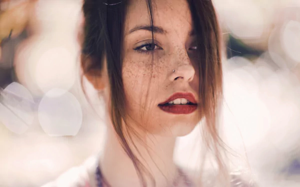 A brunette woman with freckled skin and bold red lipstick gazes confidently, set against a soft bokeh background. This HD wallpaper captures her striking features beautifully.