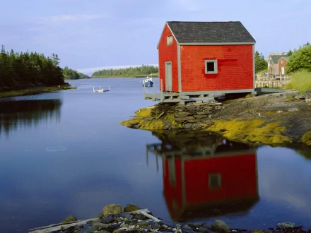 A vibrant red building reflects in calm water, surrounded by lush greenery and a serene Canadian landscape, showcasing man-made beauty against a tranquil backdrop.