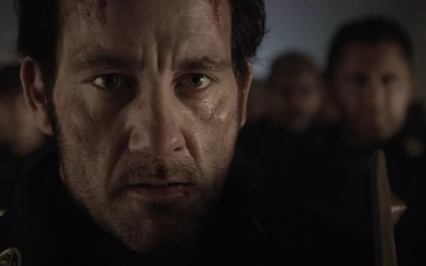 Close-up of a grim, battle-worn man in medieval armor with blurred soldiers behind him — HD movie desktop wallpaper from Last Knights.