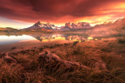 Vivid sunset over the Torres del Paine mountains in Patagonia, Chile, with clouds reflecting on a calm lake surrounded by grass and natural landscape.