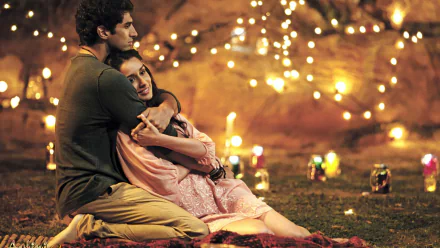 A romantic moment from the movie Aashiqui 2 featuring a couple hugging outdoors, surrounded by glowing lights, in a HD desktop wallpaper background.