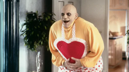 A character from *Addams Family Values* humorously poses in a heart-shaped apron, showcasing the film's distinct dark comedy and quirky charm in this HD desktop wallpaper.