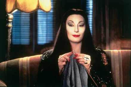 Morticia Addams from the 1991 movie The Addams Family, elegantly seated and knitting, exudes her signature dark charm in this HD desktop wallpaper.