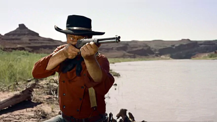 HD PC desktop wallpaper featuring John Wayne in a classic Western scene from the movie The Searchers, aiming a rifle by a river with desert hills in the background.