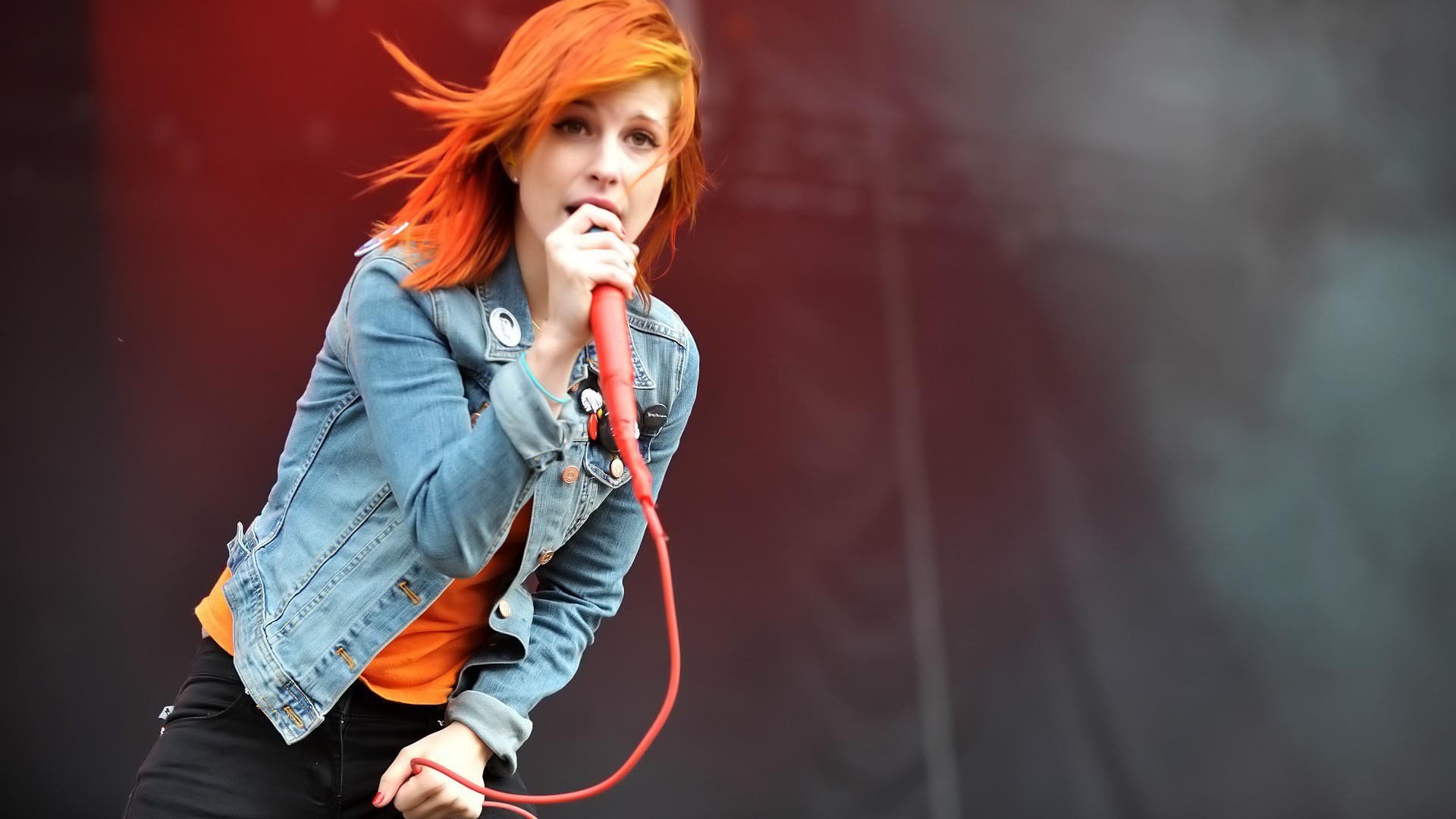 Vibrant HD PC desktop wallpaper: female rock singer with bright orange hair gripping a red microphone on a smoky stage — music background.