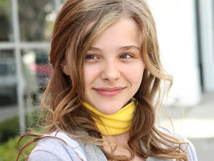 A vibrant HD desktop wallpaper featuring Chloë Grace Moretz, showcasing her smiling with soft, wavy hair and a yellow scarf, set against a blurred outdoor background.