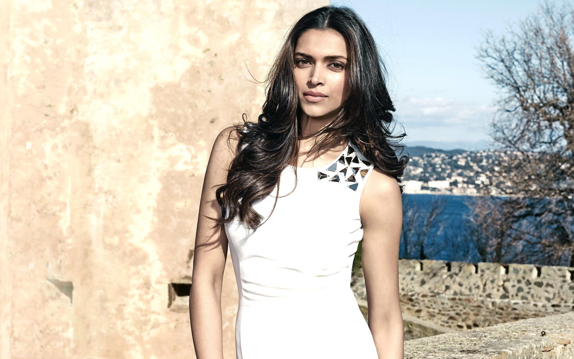 Deepika Padukone poses elegantly in a stylish white dress, framed by a scenic background of water and trees, showcasing her grace as an Indian actress and model in this HD wallpaper.