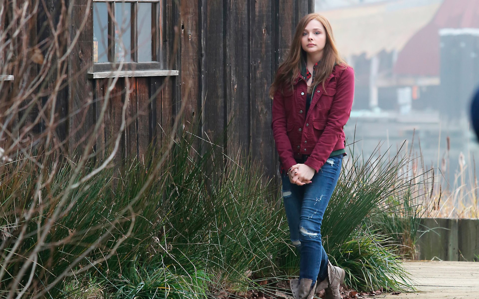 Chloë Grace Moretz stands pensively outside a rustic wooden building, surrounded by tall grass, captured in this striking HD PC desktop wallpaper.