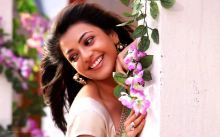 HD desktop wallpaper featuring celebrity Kajal Aggarwal, smiling and leaning against a wall adorned with pink flowers.