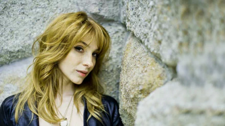 HD PC desktop wallpaper of a woman with shoulder-length blonde hair leaning against a stone wall, wearing a leather jacket and gazing thoughtfully to the side.