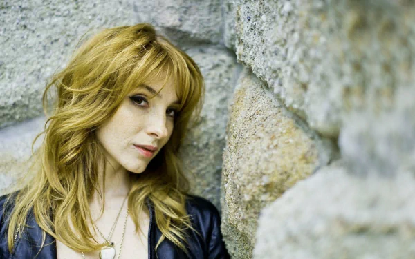 HD PC desktop wallpaper of a woman with shoulder-length blonde hair leaning against a stone wall, wearing a leather jacket and gazing thoughtfully to the side.