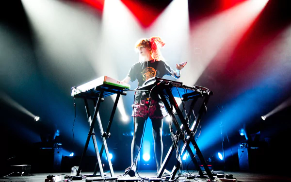 HD PC wallpaper of Grimes performing live on stage, surrounded by keyboards and colorful stage lights, capturing an energetic music moment.