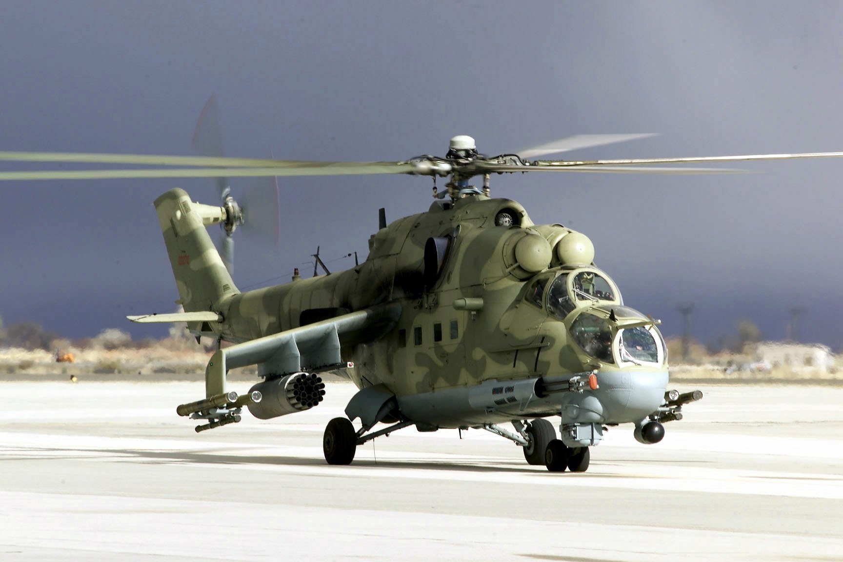 HD desktop wallpaper showing a military Mil Mi-24 helicopter on a runway with rotors spinning against a muted sky background.