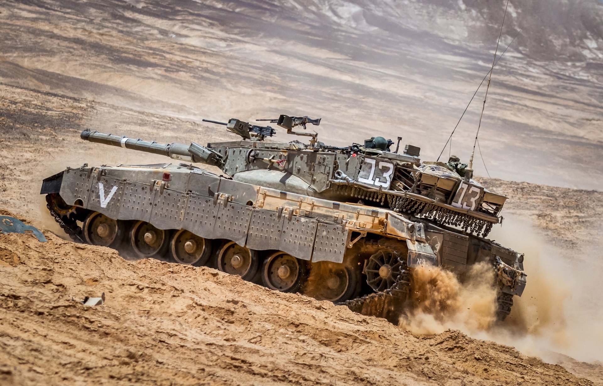 HD PC desktop wallpaper: a Merkava main battle tank kicking up dust as it maneuvers across a rugged desert landscape, turret and antennas clearly visible.