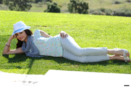 A HD desktop wallpaper featuring Selena Gomez lounging on green grass, wearing a white hat and a light outfit, exuding a relaxed and cheerful vibe surrounded by nature.
