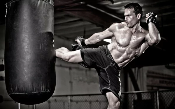 A muscular man executes a powerful kick on a heavy punching bag in a gym. This HD wallpaper and background evokes themes of fitness and sports.
