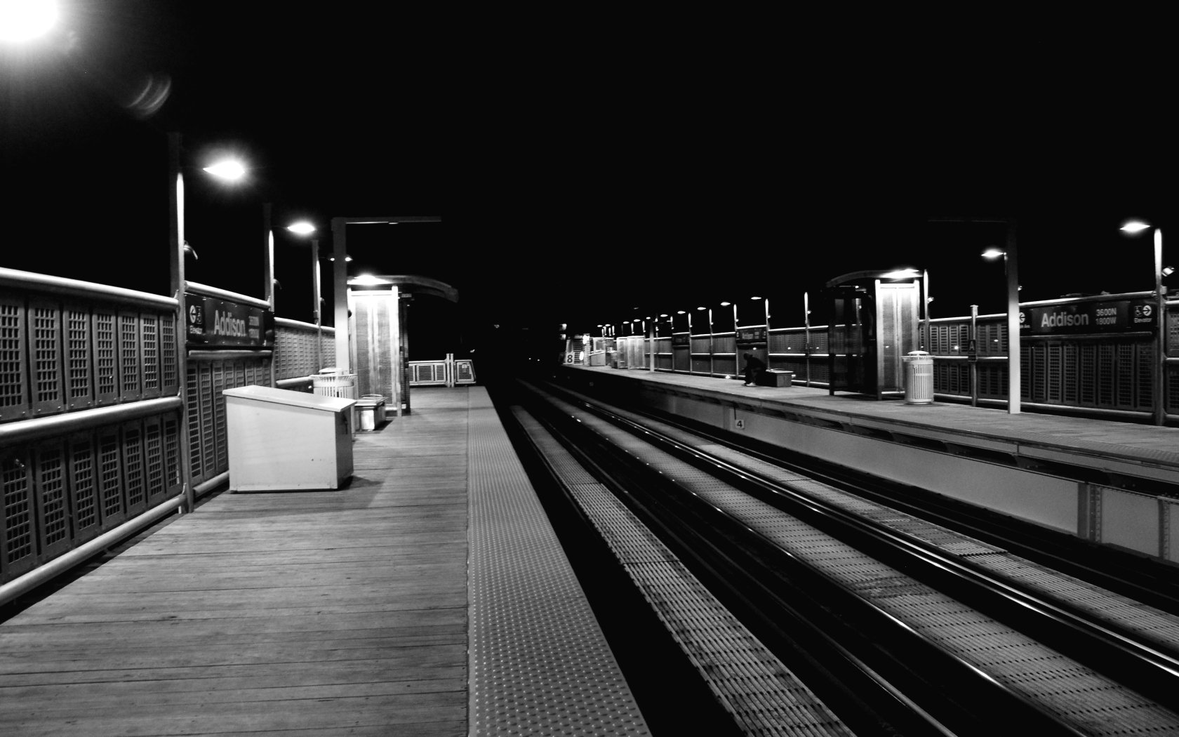 A black-and-white HD desktop wallpaper depicting a quiet, empty railroad station at night, featuring illuminated platforms and parallel train tracks.