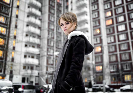 A woman stands confidently in a winter scene, wearing a black coat with a white collar, set against a modern apartment building backdrop, captured as a high-definition desktop wallpaper.