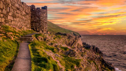 HD desktop wallpaper showing the man-made Peel Castle on a grassy cliffside with a vibrant sunset over the sea in the background.