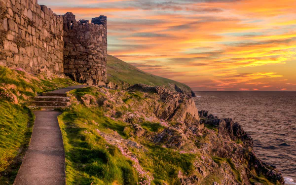 HD desktop wallpaper showing the man-made Peel Castle on a grassy cliffside with a vibrant sunset over the sea in the background.