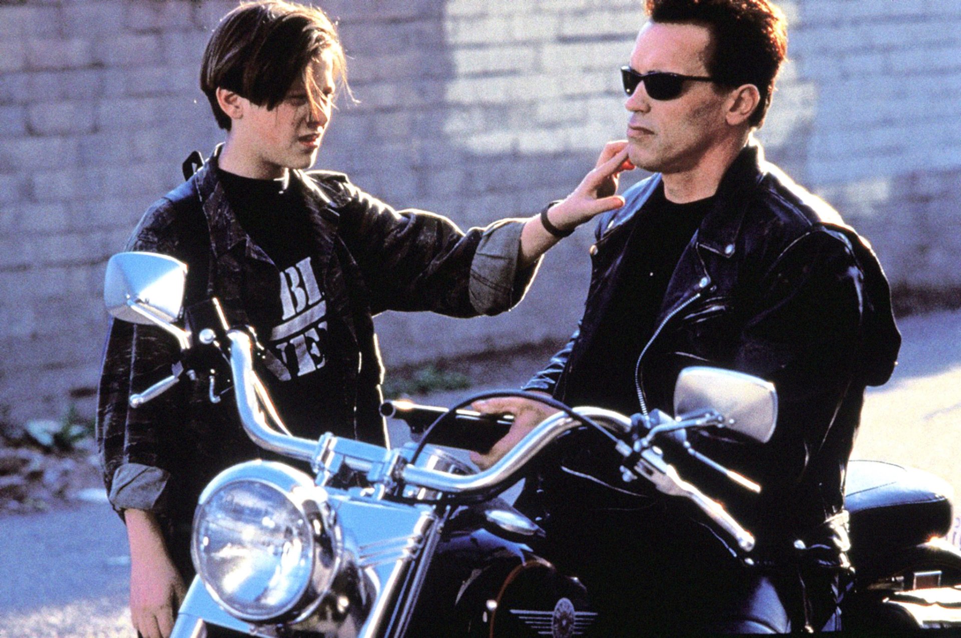 A scene from *Terminator 2: Judgment Day* featuring Arnold Schwarzenegger as the Terminator and Edward Furlong as John Connor, showcasing their bond on a motorcycle.