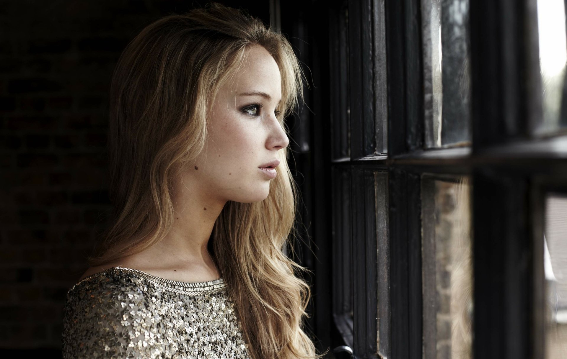 HD desktop wallpaper featuring American blonde actress Jennifer Lawrence gazing thoughtfully out of a window with soft natural light highlighting her profile.