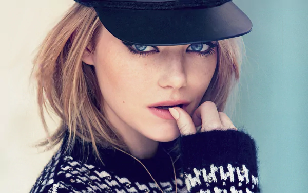 HD desktop wallpaper featuring celebrity Emma Stone. She is wearing a black cap and a patterned sweater, gazing intently at the camera with a hand near her mouth.