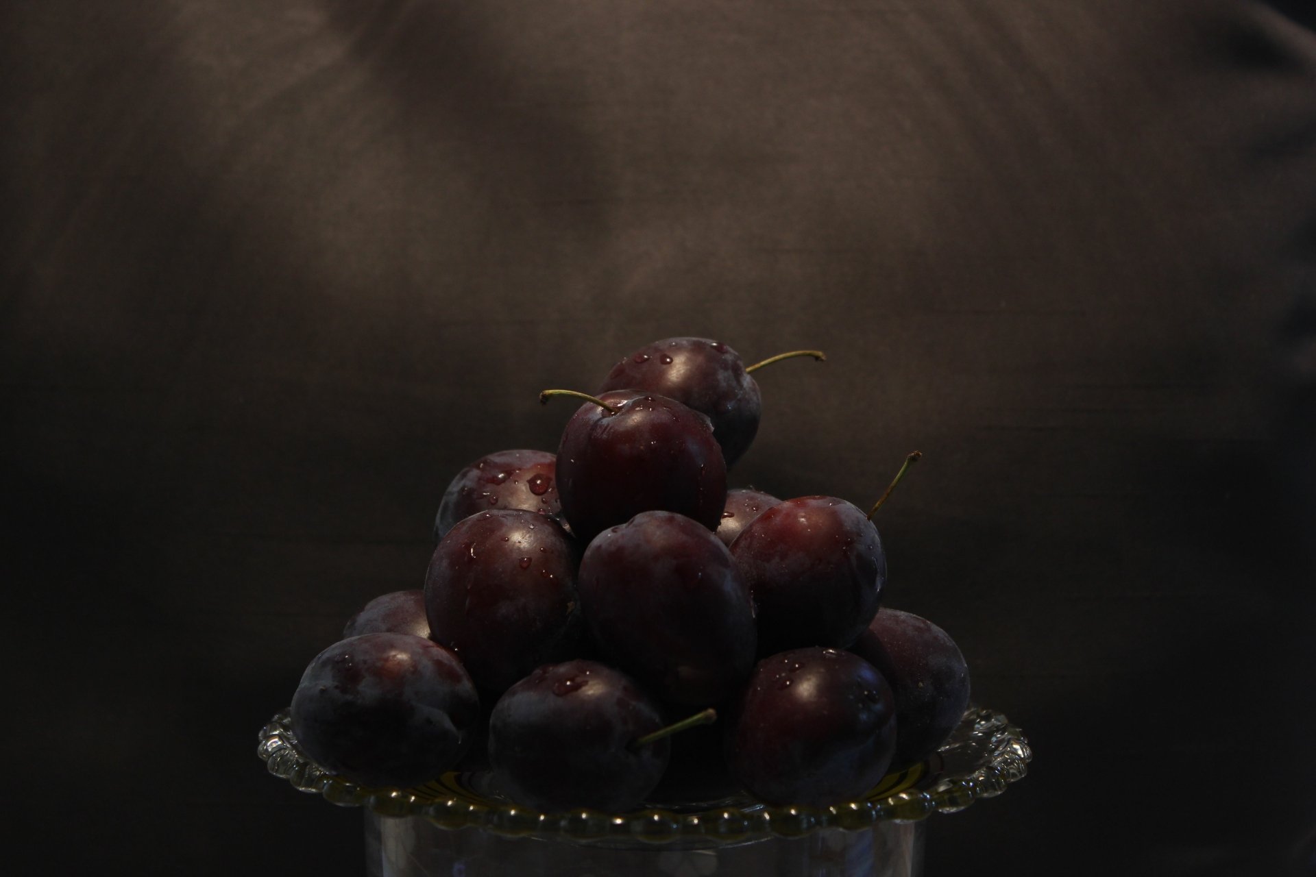 5K Ultra HD PC desktop wallpaper: food still-life of glossy dark plums piled on a glass plate, moody spotlight and deep-shadow background.