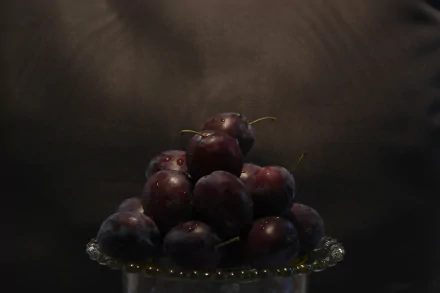 5K Ultra HD PC desktop wallpaper: food still-life of glossy dark plums piled on a glass plate, moody spotlight and deep-shadow background.