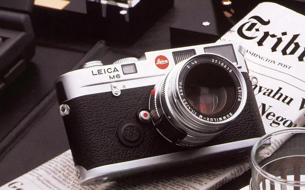 Leica M6 rangefinder on a desk beside a newspaper and glass — HD PC desktop wallpaper background showcasing a classic man-made camera