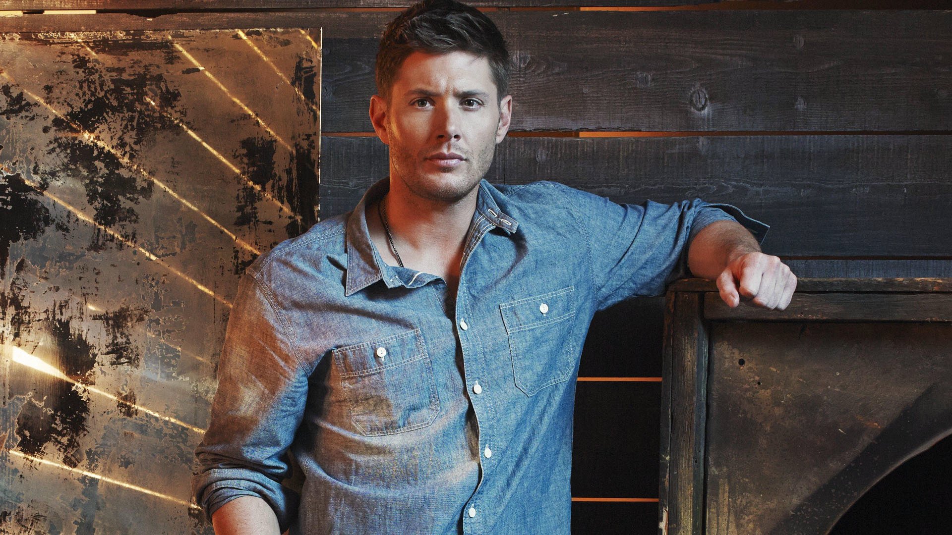 A high-definition desktop wallpaper featuring a character from the TV show Supernatural, portrayed in a casual denim shirt against a rustic background.