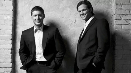Black and white image of Jared Padalecki and Jensen Ackles as Sam and Dean Winchester from the TV show Supernatural, both dressed in suits against a textured wall backdrop.