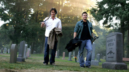 Two characters walk through a misty graveyard, dressed casually and carrying jackets, capturing a moment from the TV show Supernatural. The scene is atmospheric and engaging.