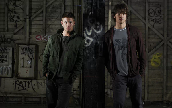 Two characters from the TV show Supernatural stand confidently in an urban setting with graffiti on the walls, showcasing a moody atmosphere. HD desktop wallpaper background.