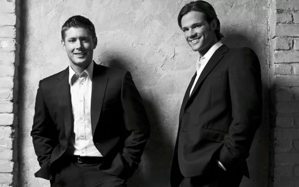 Black and white image of Jared Padalecki and Jensen Ackles as Sam and Dean Winchester from the TV show Supernatural, both dressed in suits against a textured wall backdrop.