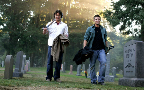 Two characters walk through a misty graveyard, dressed casually and carrying jackets, capturing a moment from the TV show Supernatural. The scene is atmospheric and engaging.