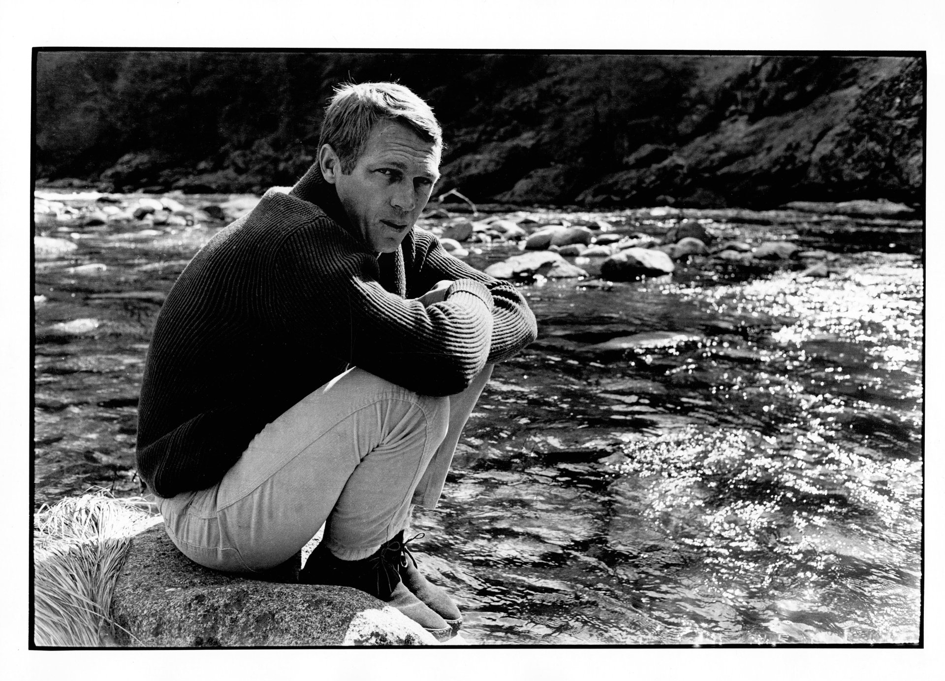 Black-and-white 4K Ultra HD PC desktop wallpaper of a classic Hollywood celebrity seated on a riverside rock, wearing a sweater and light trousers, gazing thoughtfully at the water.