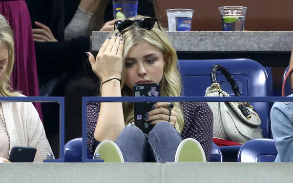 Chloë Grace Moretz sits in a stadium, focused on her phone while adjusting her sunglasses, surrounded by fans enjoying the event. This image serves as a vibrant 4K Ultra HD wallpaper.