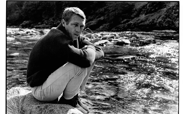 Black-and-white 4K Ultra HD PC desktop wallpaper of a classic Hollywood celebrity seated on a riverside rock, wearing a sweater and light trousers, gazing thoughtfully at the water.
