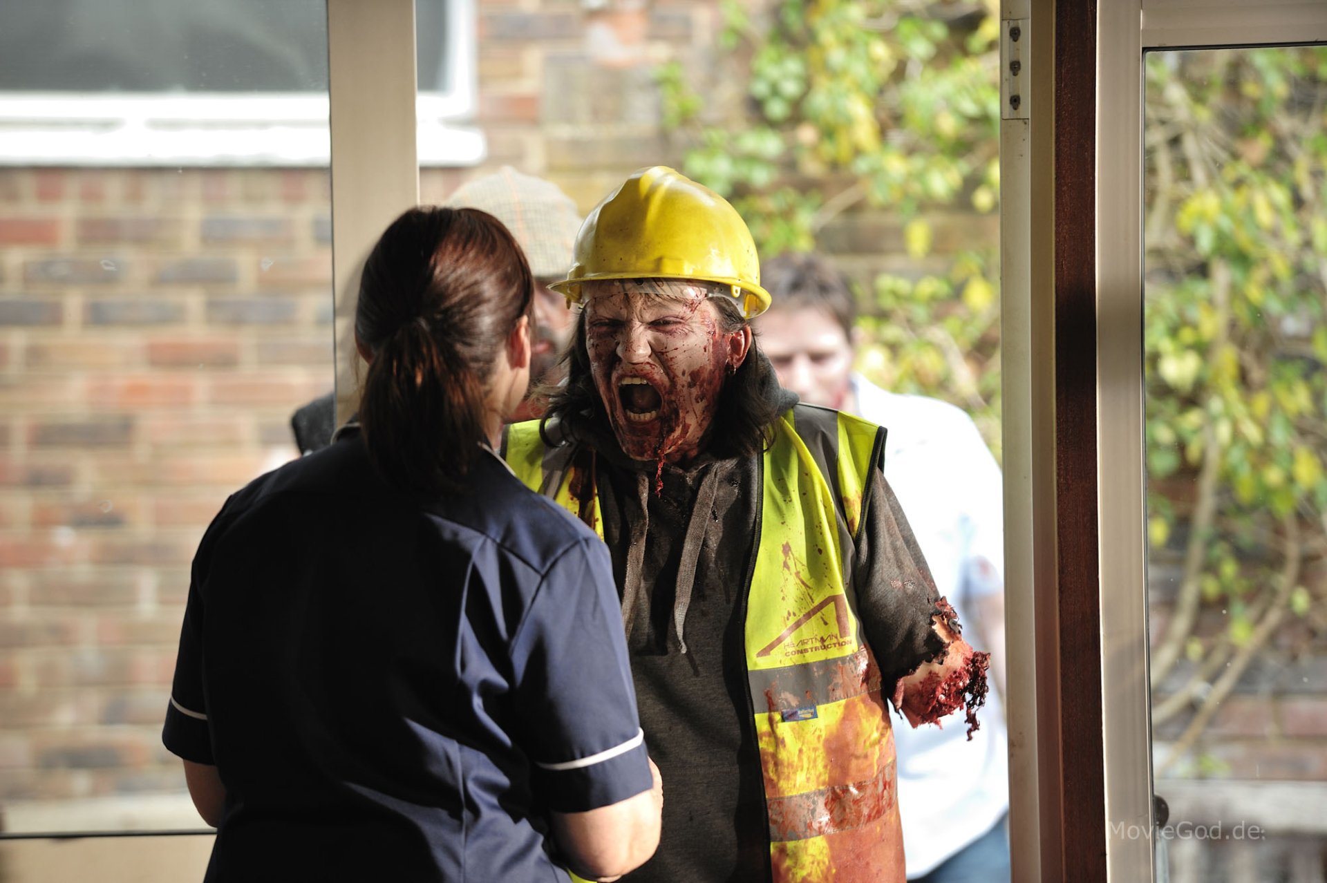HD PC desktop wallpaper and background: still from the movie Cockneys vs Zombies — a bloodied construction-worker zombie in a hi‑vis vest and hard hat lunges at a nurse in a doorway.