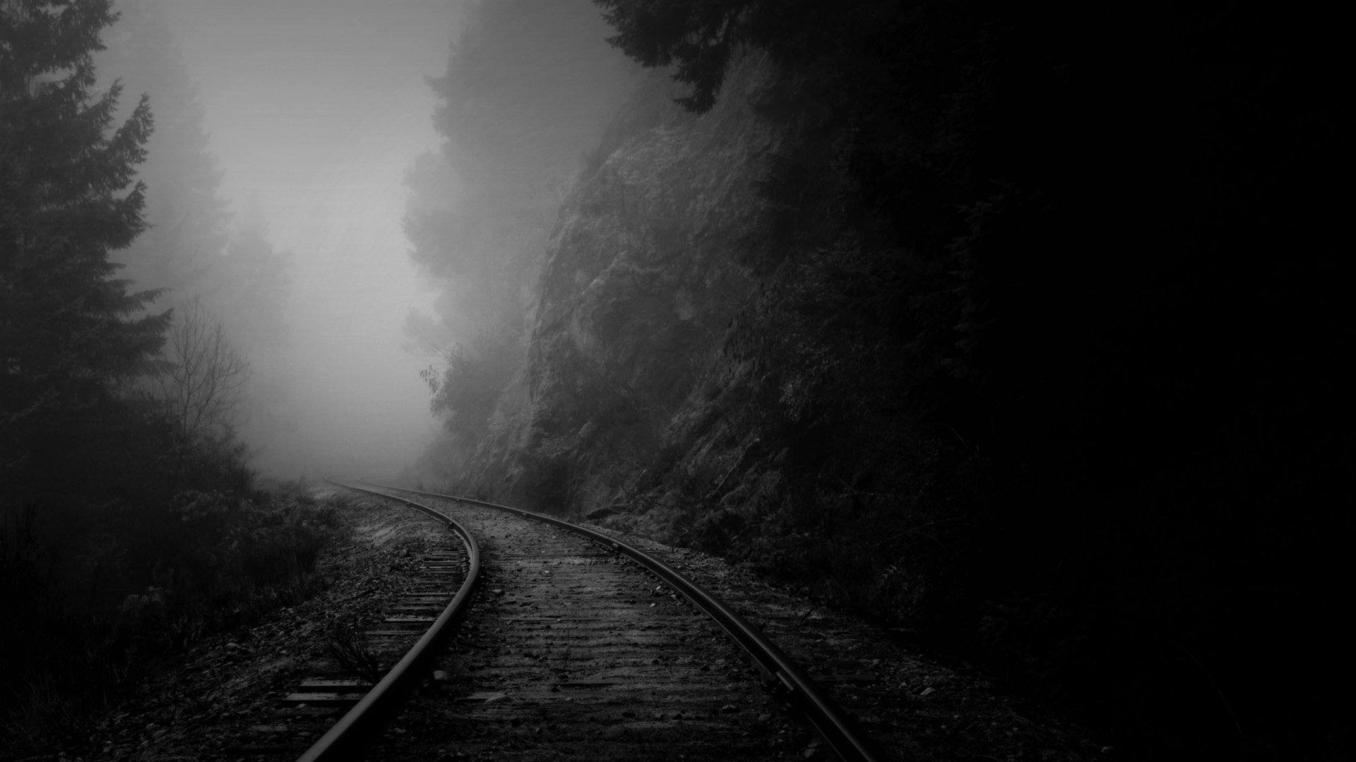 HD desktop wallpaper of a misty railroad curving alongside a rocky cliff, capturing a moody, atmospheric scene of man-made railway tracks disappearing into fog.