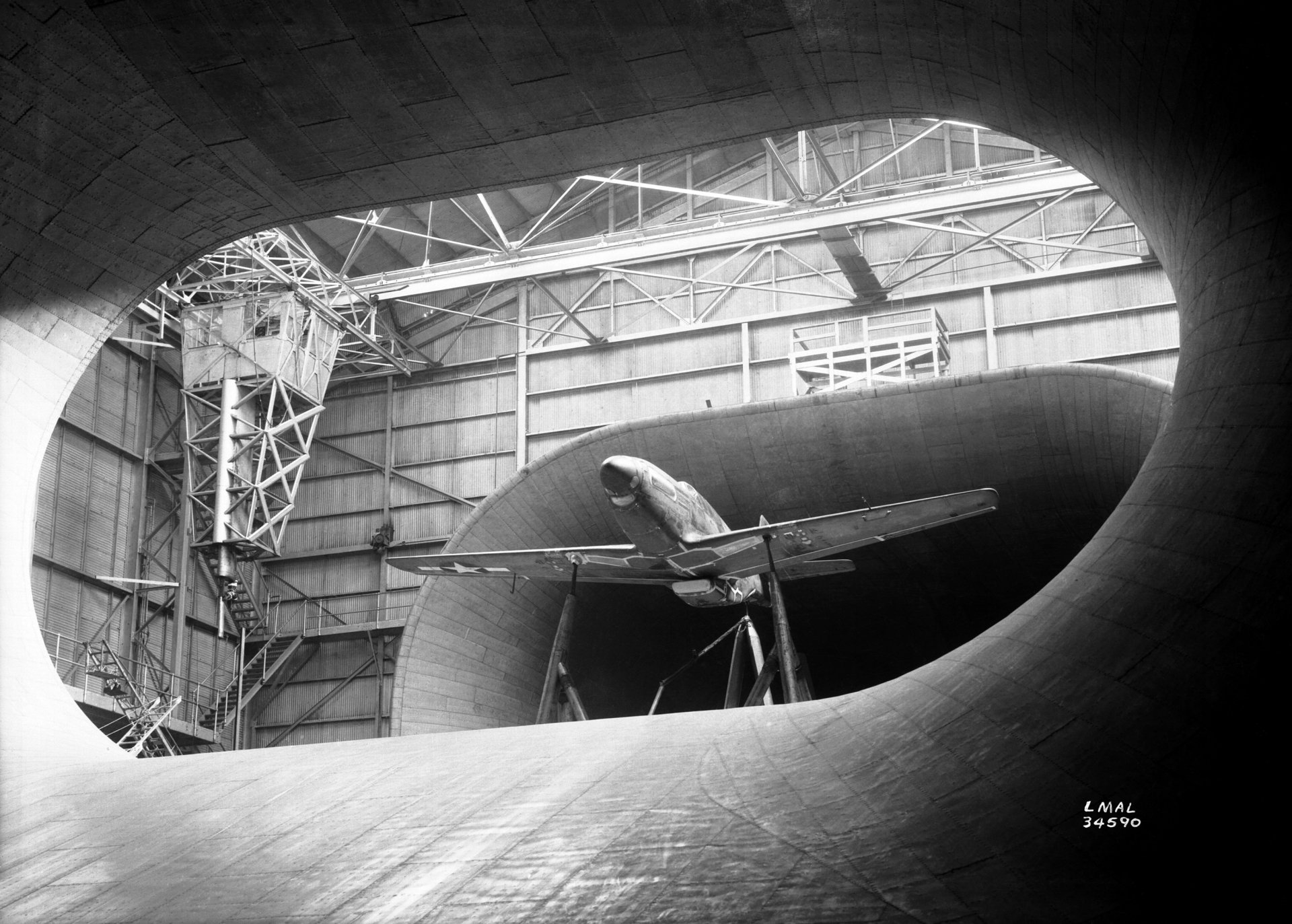 Black-and-white 2K Quad HD desktop wallpaper showing a man-made North American P-51 Mustang aircraft inside a NASA wind tunnel, seen through the tunnel's circular opening.