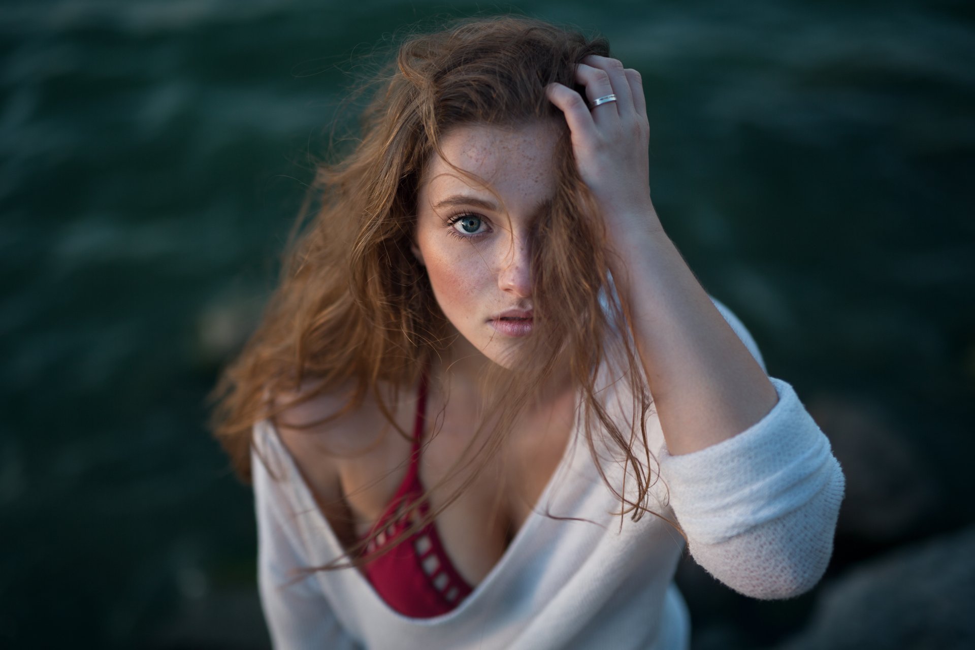 A stunning brunette model with freckles and blue eyes gazes intently at the camera, framed by a blurred natural backdrop in this captivating 4K Ultra HD wallpaper.